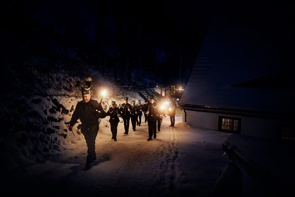 Eine Gruppe von Menschen in Uniform geht nachts mit Fackeln an einem verschneiten Weg entlang und beleuchtet dabei die Umgebung in der Nähe des Bergbaumuseums. Rechts ist ein Gebäude mit beleuchteten Fenstern zu sehen, und im dunklen Hintergrund zeichnen sich die Silhouetten von Bäumen ab.