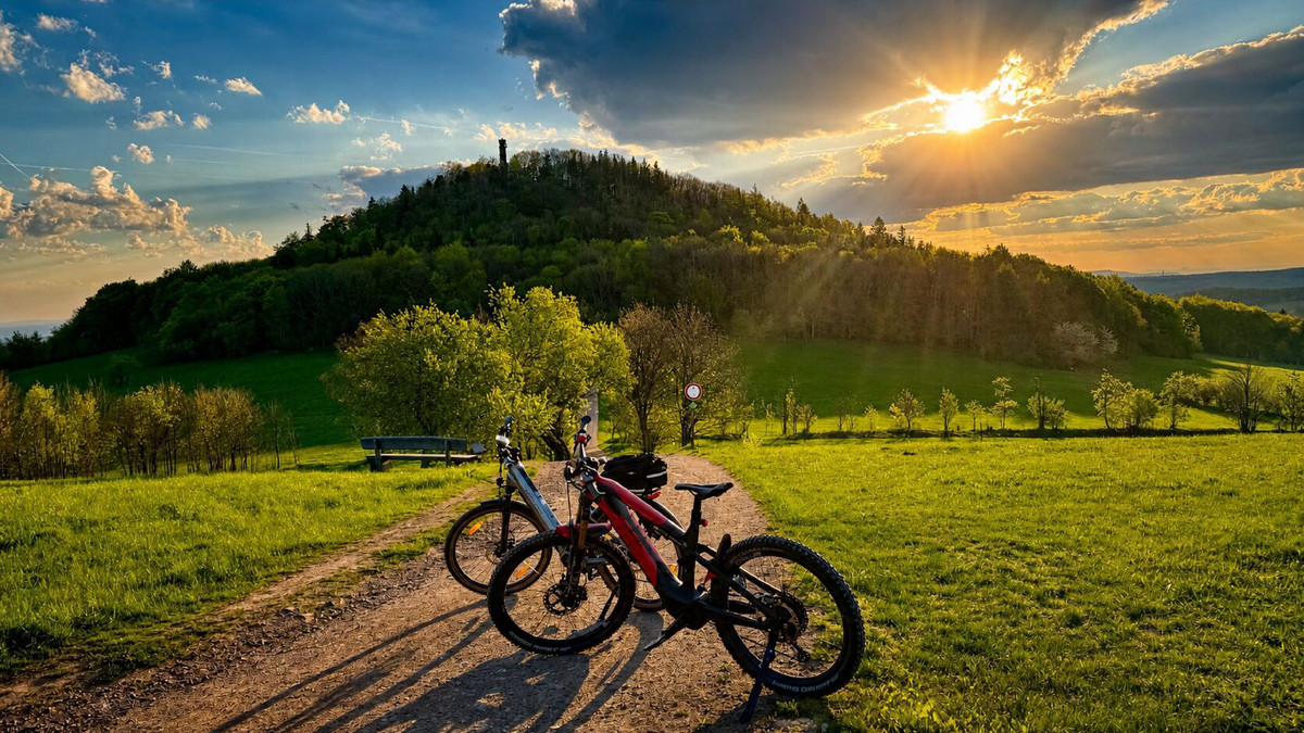 Zwei Fahrräder stehen auf einem Feldweg neben einer Bank und bieten den Blick auf den Geisingberg in der Nähe des Gasthofs Bärenfels unter einem stimmungsvollen Himmel bei Sonnenaufgang. Bäume und üppige Felder umgeben den Hügel und schaffen eine friedliche Landschaft, die die Gäste des Naturhotels Bärenfels genießen können.