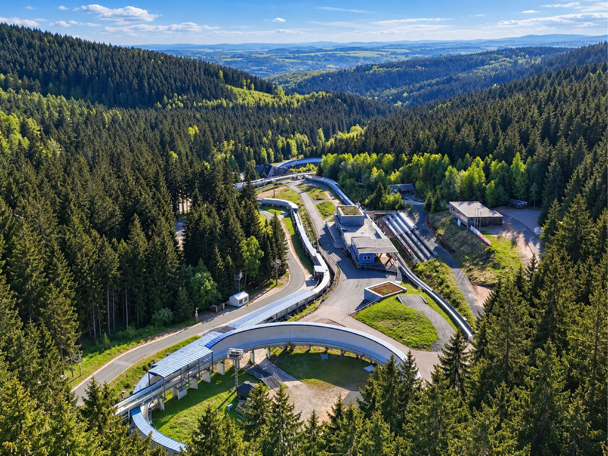 Eine kurvenreiche Bob- oder Rodelbahn schlängelt sich durch einen dichten, grünen Wald in einer hügeligen Landschaft – ideal für einen Aktivurlaub. Unter strahlend blauem Himmel, mit Blick auf die fernen Berge, liegen Gebäude und Wege verstreut rund um die Anlage.