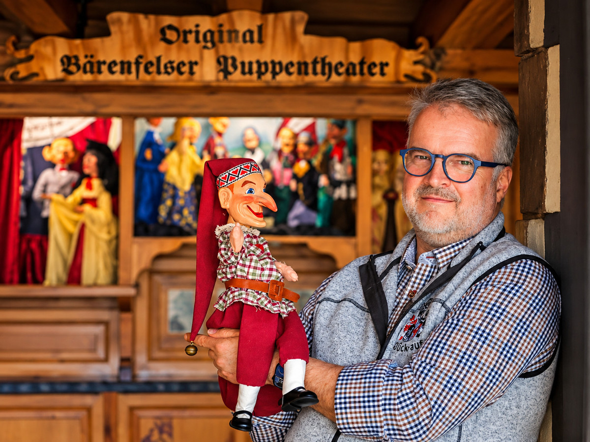 Ein Mann mit Brille hält eine traditionelle Marionette vor einer Bühne mit der Aufschrift „Original Bärenfelser Puppentheater“ im Gasthof Bärenfels in die Kamera, während im Hintergrund mehrere bunte Marionetten zu sehen sind.