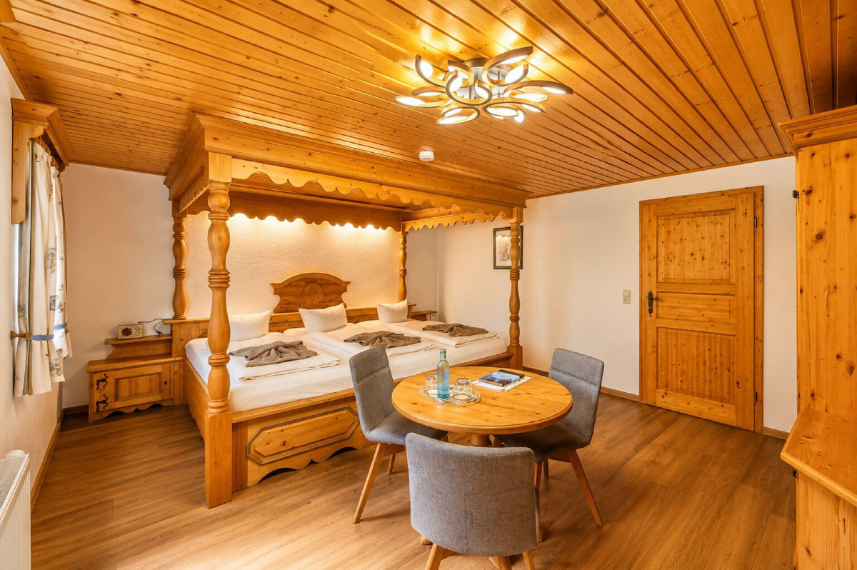 Gemütliches Familienzimmer aus Holz im Naturhotel Gasthof Bärenfels mit einem extragroßen Himmelbett, einem runden Tisch, einer modernen Deckenleuchte und viel Tageslicht. Mineralwasser und Gläser stehen für Sie bereit – genießen Sie die warme, einladende Atmosphäre in diesem charmanten Naturhotel.