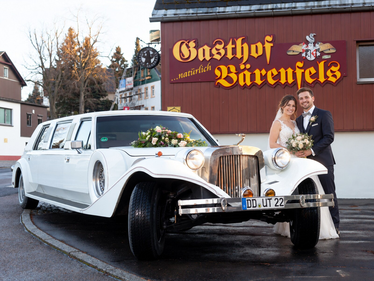 Ein Brautpaar steht lächelnd neben einer weißen, mit Blumen geschmückten Oldtimer-Limousine, die vor dem Gasthof Bärenfels geparkt ist – und feiert seine Hochzeit im malerischen Erzgebirge.