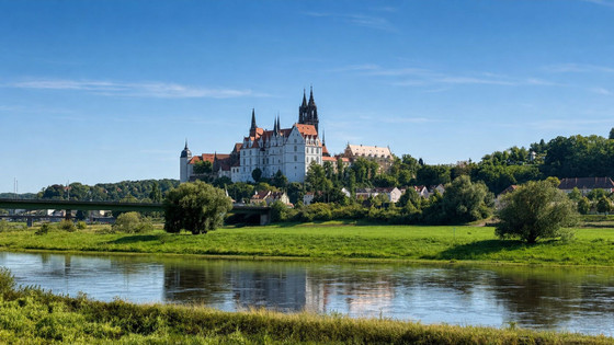 Auf einem Hügel oberhalb des Elbtals thront eine große, historische Burg mit Türmen, von der aus man auf eine kleine Stadt und die Weinberge des berühmten Weinbaugebiets der Region blickt. Im Vordergrund fließt ein spiegelglatter Fluss unter einem blauen Himmel mit vereinzelten Wolken.