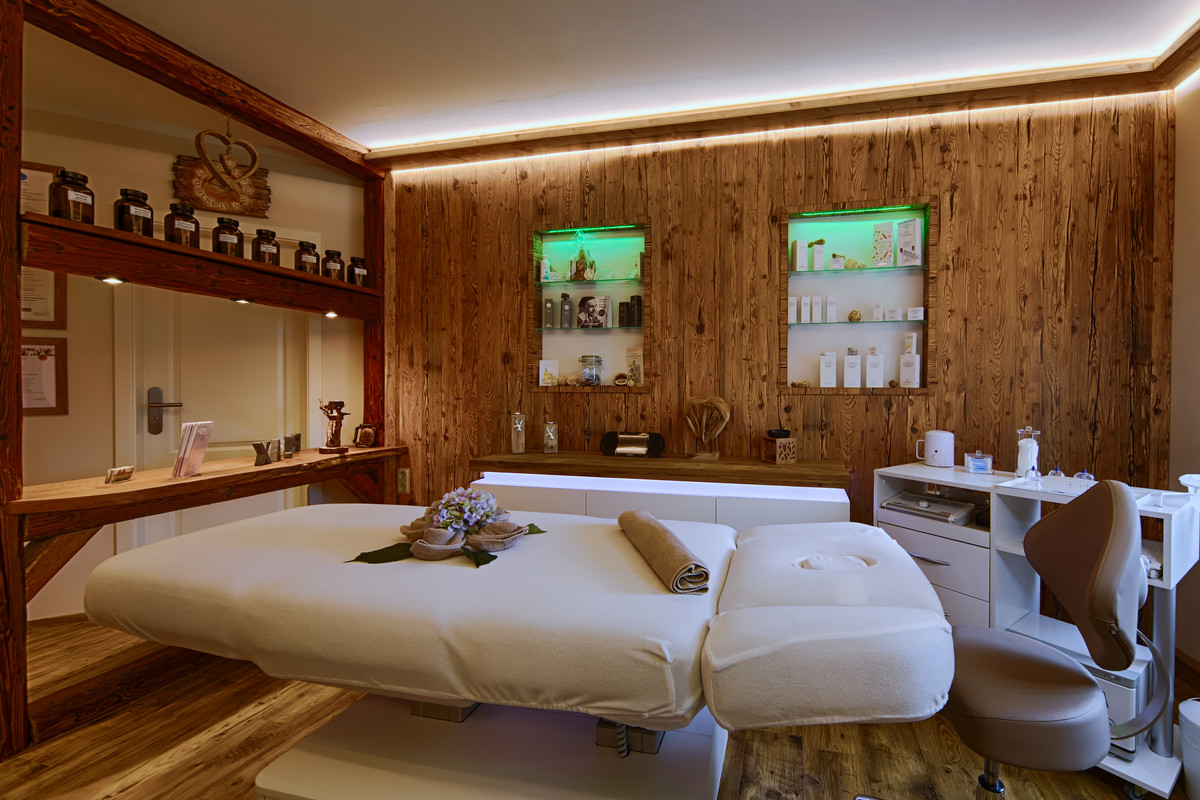 Ein gemütlicher Spa-Behandlungsraum mit einer Massageliege, Holzwänden, Regalen mit Gläsern und Hautpflegeprodukten, einem Stuhl und sanfter Stimmungsbeleuchtung, die eine entspannte Atmosphäre schafft.