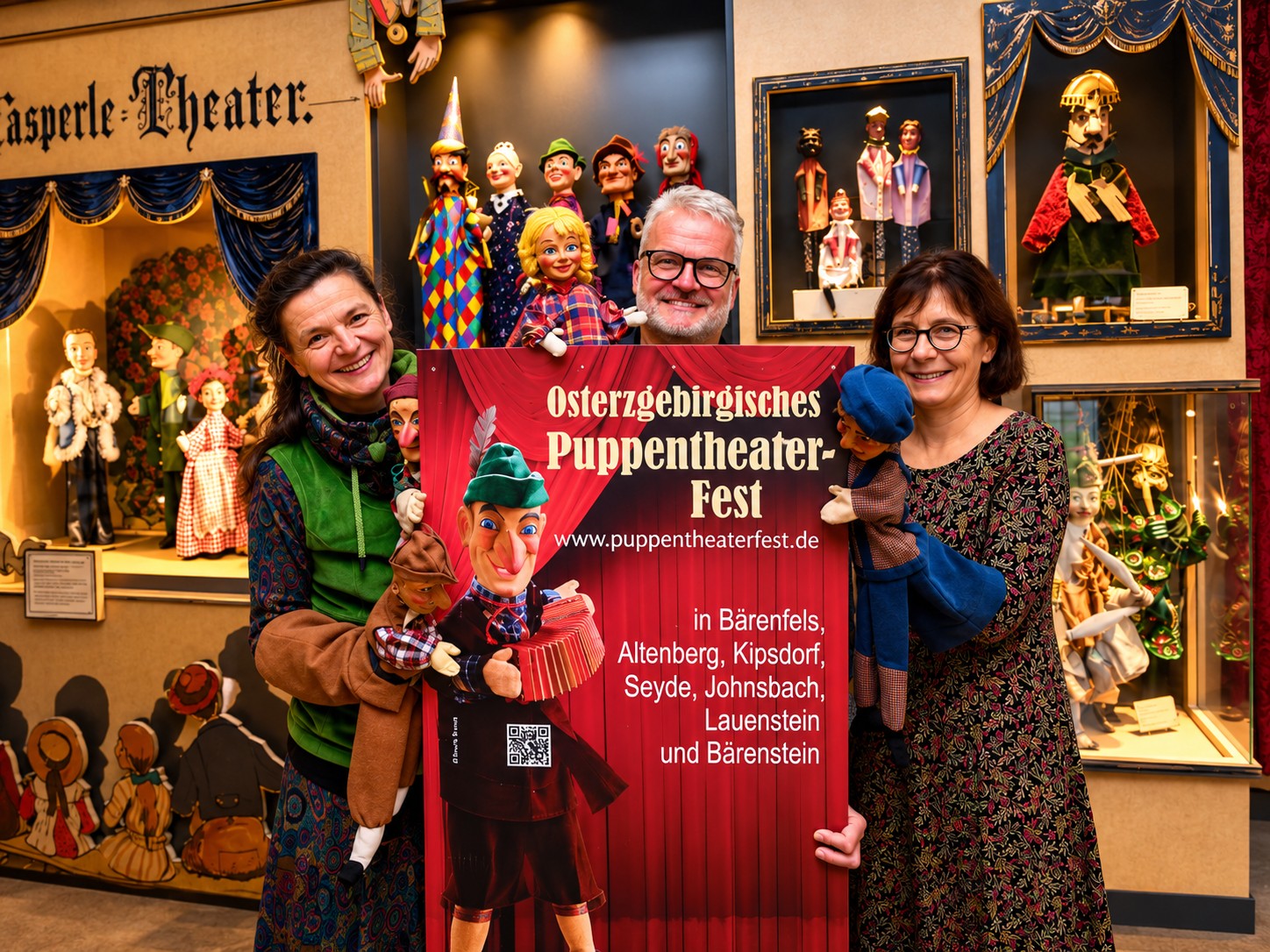 Drei lächelnde Erwachsene stehen in einem Puppentheater und halten Handpuppen sowie ein großes Plakat in den Händen, das für das Osterzgebirgische Puppentheater-Fest wirbt. Puppenausstellungen, detailreiche Dekorationen und Informationen zu Veranstaltungen prägen den farbenfrohen Hintergrund.