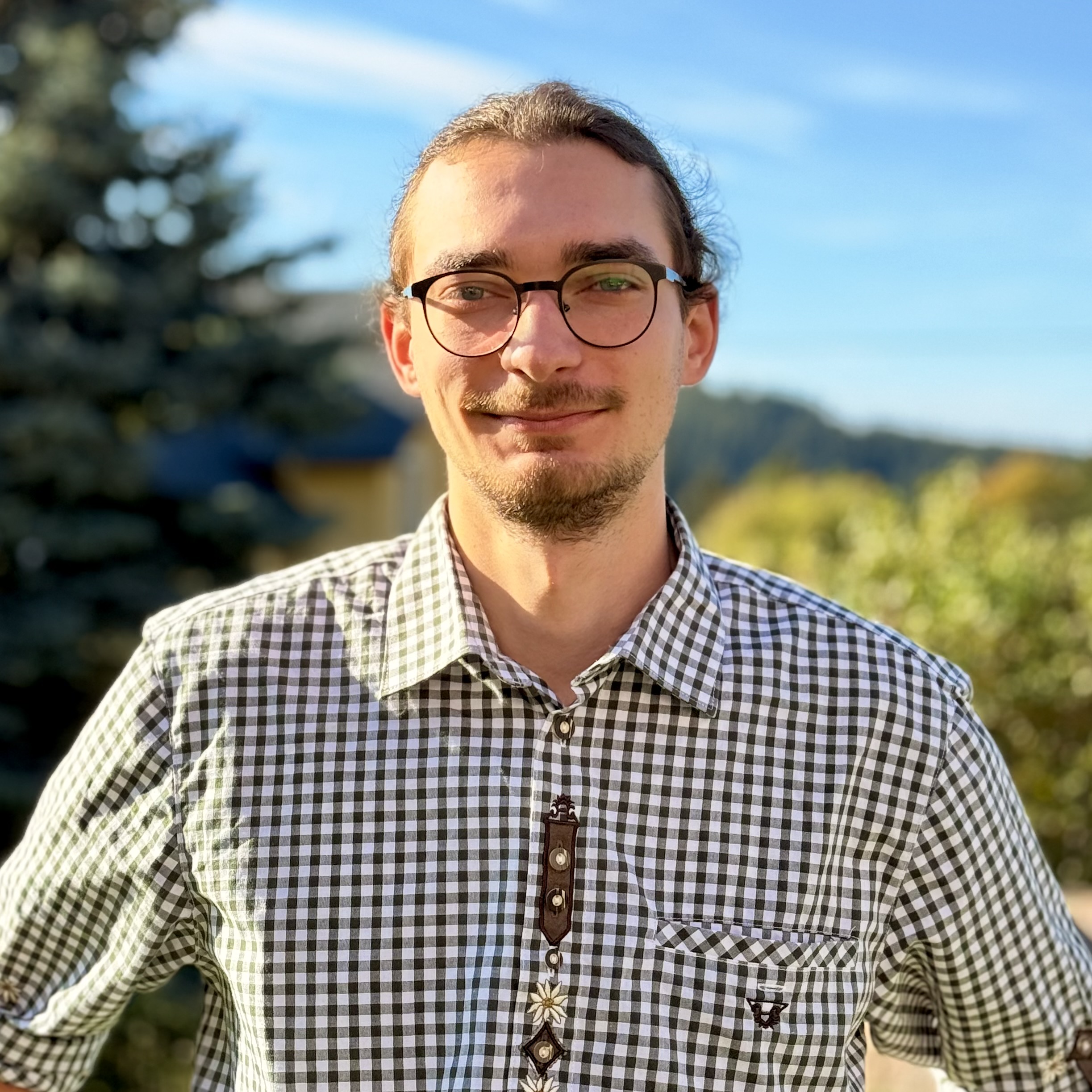 Ein junger Mann mit Brille und Spitzbart, der draußen ein grün-weiß kariertes Hemd mit Knöpfen trägt.