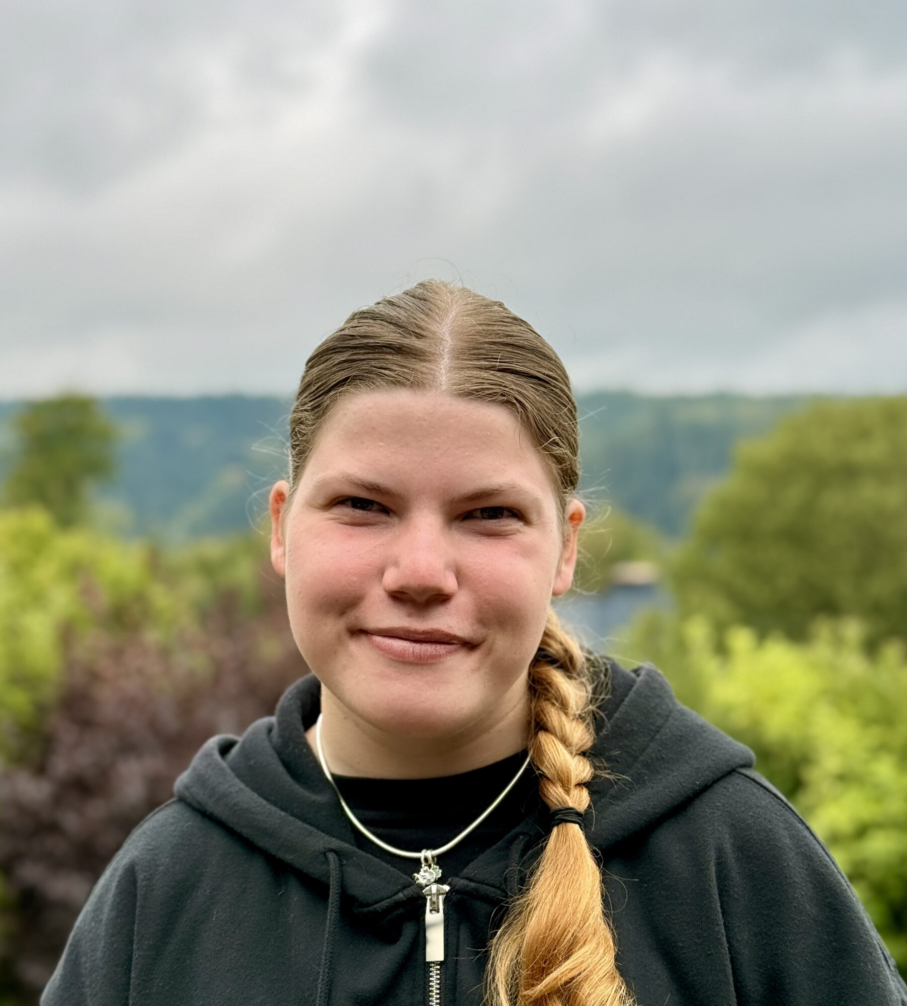 Ein junger Mensch mit einem blonden Zopf, der einen schwarzen Kapuzenpulli und eine silberne Halskette trägt, lächelt vor dem Hintergrund von Bäumen und einem bewölkten Himmel in die Kamera.