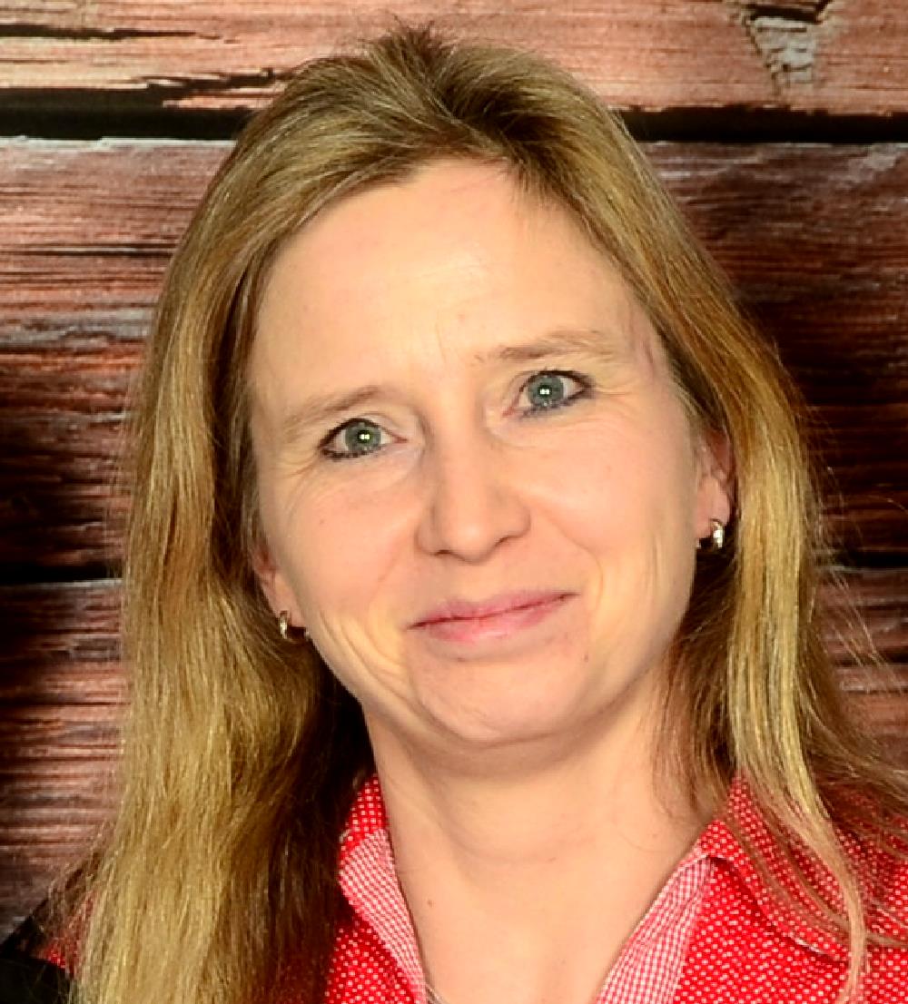 Eine Frau mit schulterlangen blonden Haaren, die vor einem Hintergrund mit Holzstruktur lächelt.