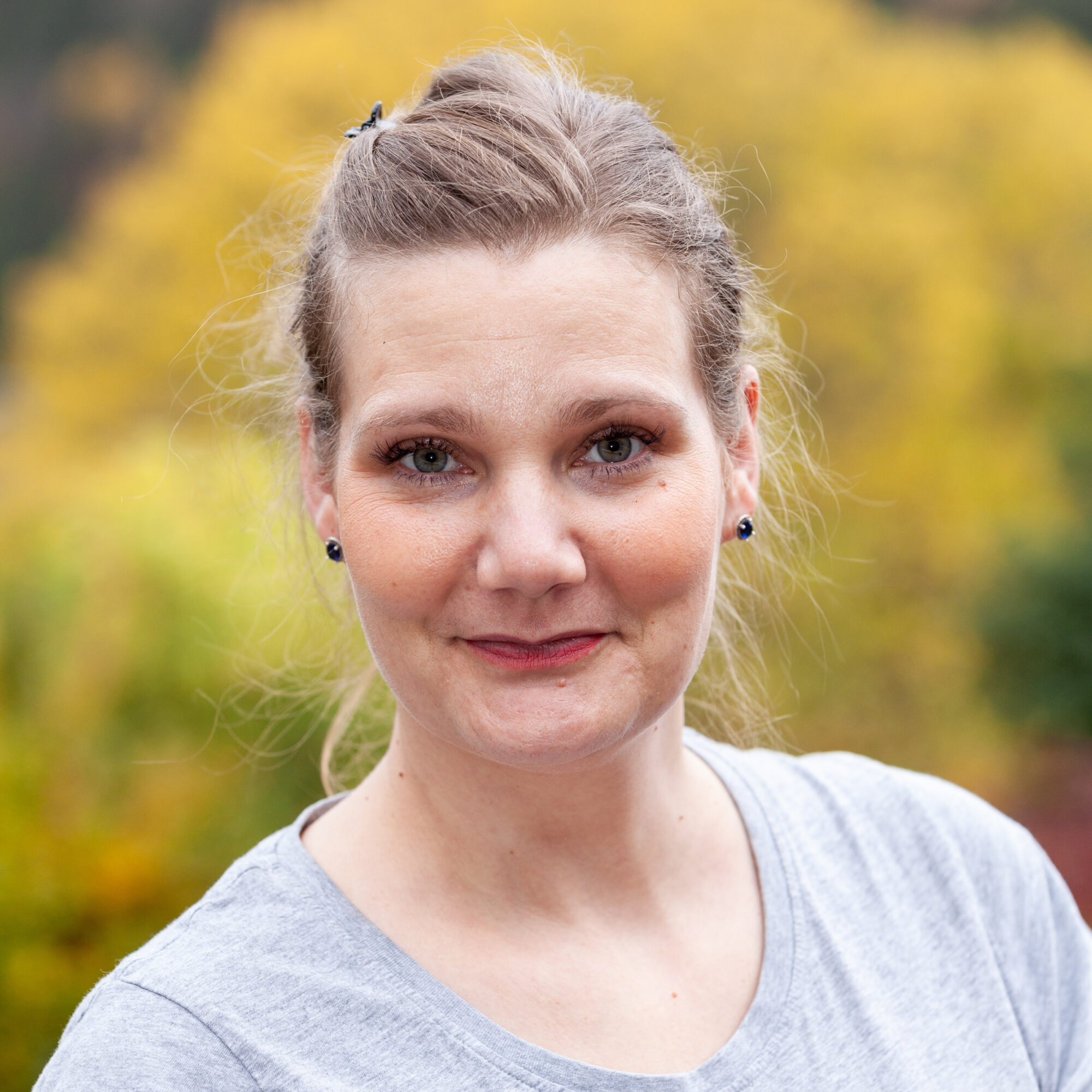 Eine Frau mit braunen, nach hinten gebundenen Haaren in einem grauen T-Shirt, die in die Kamera lächelt.