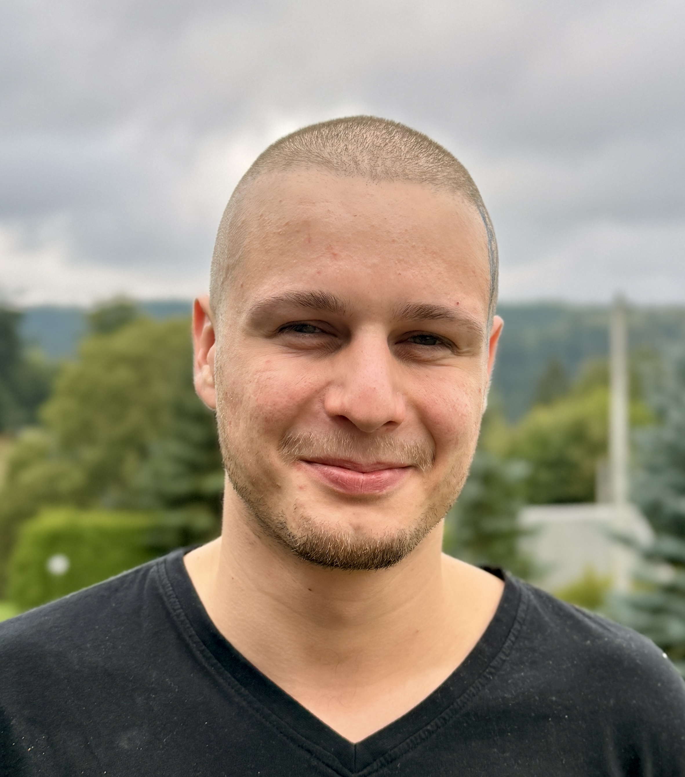 Ein junger Mann mit rasiertem Kopf und leichtem Bartstoppeln, der in die Kamera lächelt.