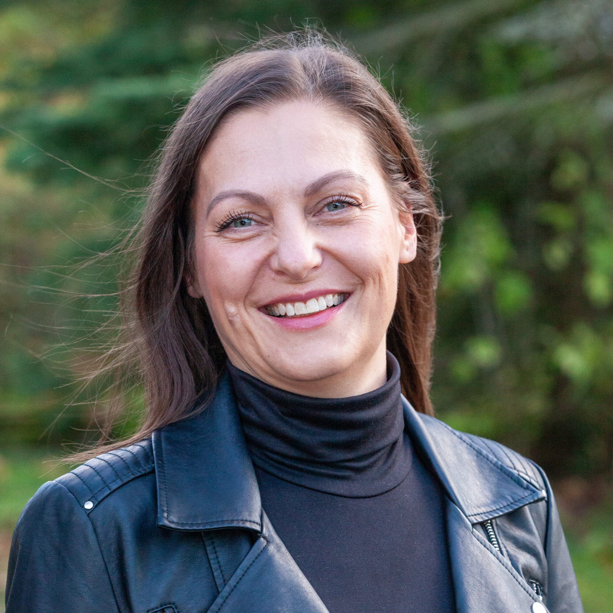 Eine lächelnde Frau mit langen braunen Haaren, die einen schwarzen Rollkragenpullover und eine schwarze Lederjacke trägt.