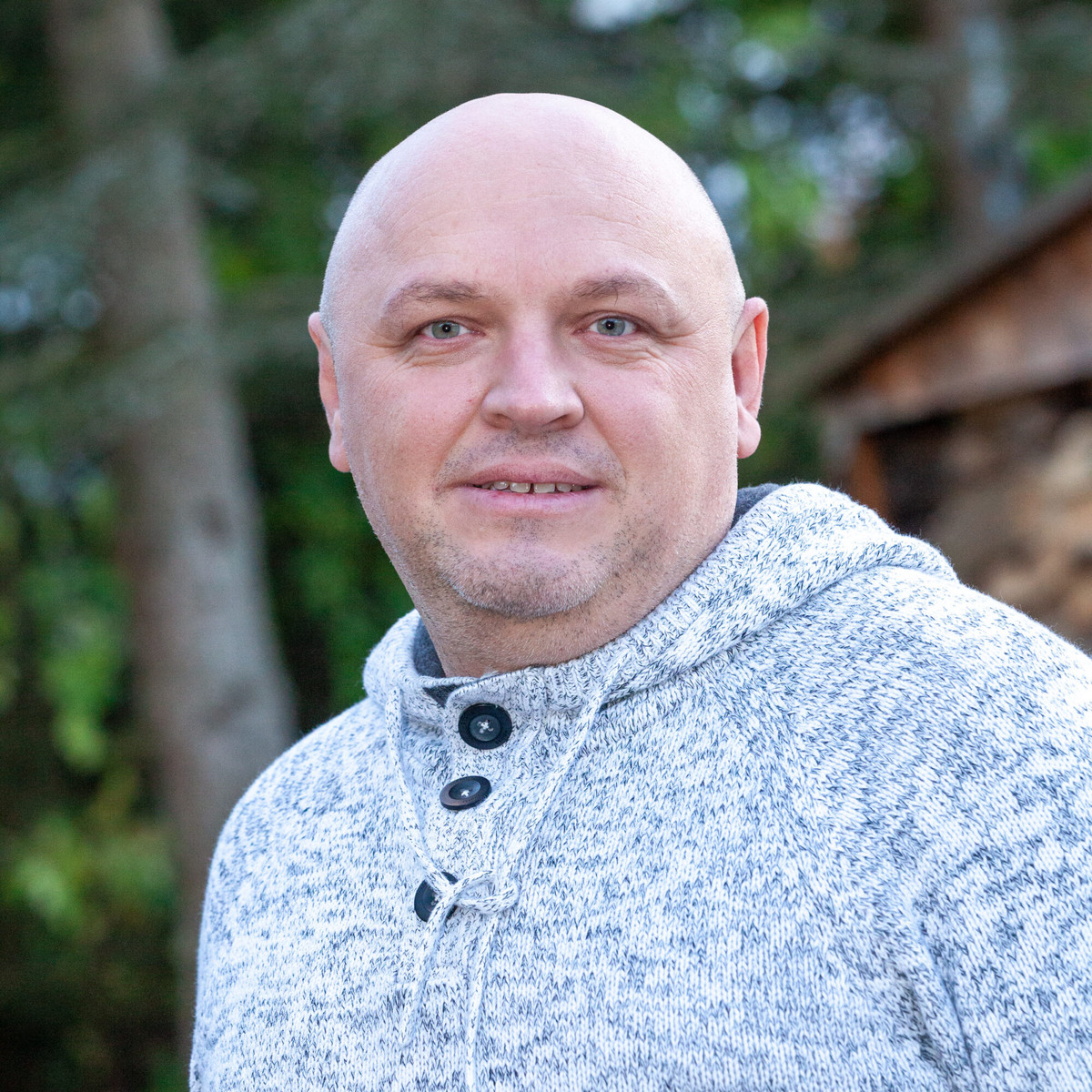 Ein Mann mittleren Alters mit rasiertem Kopf, der draußen einen grauen Kapuzenpullover trägt.