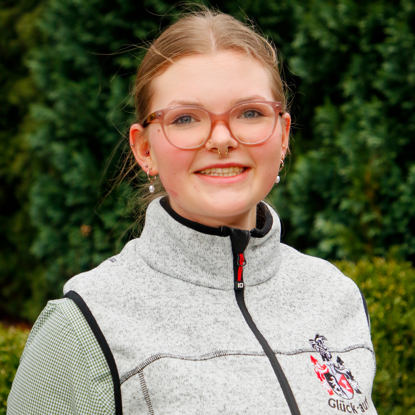 Eine junge Frau mit Brille, die eine graue Weste mit Reißverschluss trägt, auf der ein Wappen und der Schriftzug Glück-auf zu sehen sind.