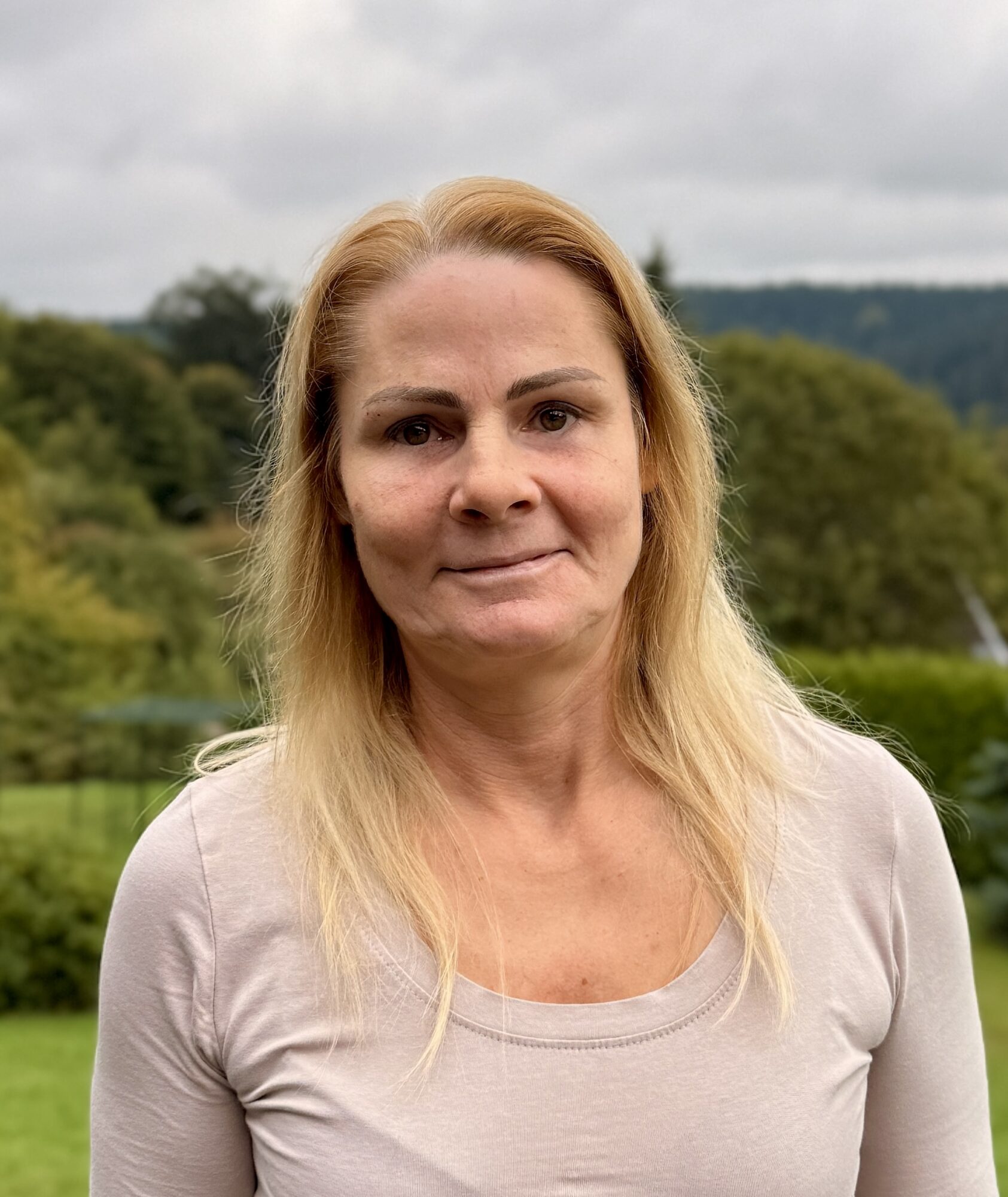 Das Porträt einer Frau mit blonden Haaren, die ein beiges Langarmhemd trägt und vor einer verschwommenen grünen Landschaft steht.